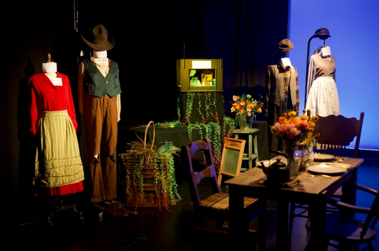 Display of the final projects done by the Drama: Production & Design students including costumes on mannequins, dioramas, and a dining table with place settings, a vase with silk flowers, a kettle, and chairs.